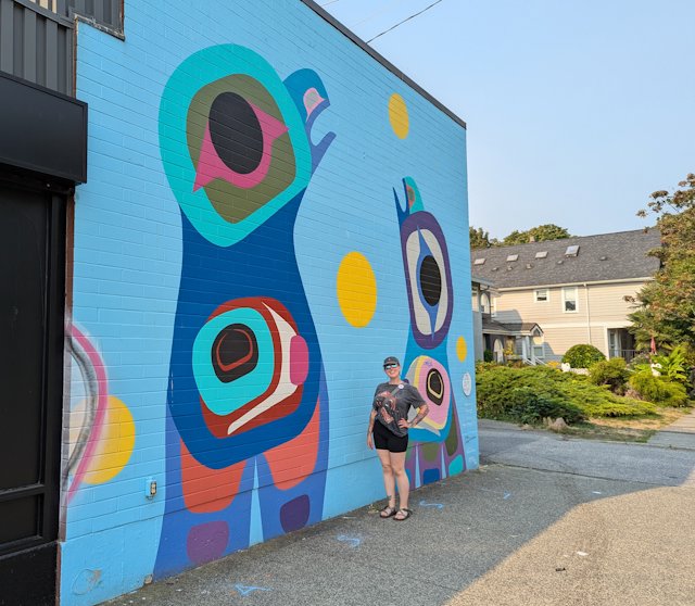 Stacey poses in front of a bright blue building with large bird totem things painted on it